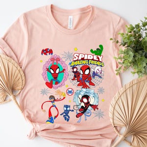 May include: A light pink t-shirt featuring a colorful graphic of Spider-Man and friends. The design includes the text "Spidey and Amazing Friends" and various cartoon illustrations of the characters. The shirt is made of soft material.