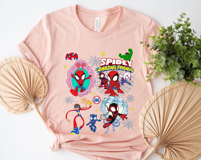May include: A light pink t-shirt featuring a colorful graphic of Spider-Man and friends. The design includes the text "Spidey and Amazing Friends" and various cartoon illustrations of the characters. The shirt is made of soft material.