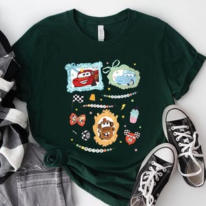 Puede incluir: Camiseta verde oscuro con personajes de dibujos animados de coches en marcos ornamentados. La camiseta incluye una bandera a cuadros, una pajarita y un gr&aacute;fico de batido. Zapatillas de lona negras con cordones blancos junto a la camiseta.