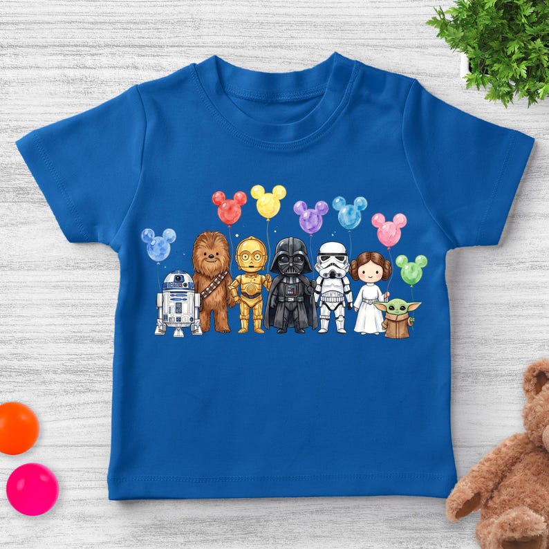 Op de afbeelding: Een blauw T-shirt met een aquarel illustratie van Star Wars-personages die Mickey Mouse-vormige ballonnen vasthouden. De personages zijn R2-D2, Chewbacca, C-3PO, Darth Vader, een Stormtrooper, Princess Leia en Grogu.