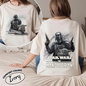 May include: Ivory-colored t-shirt featuring a graphic of The Mandalorian and Grogu from Star Wars. The front shows the characters on a ship, while the back displays them with blasters. The text "STAR WARS THE MANDALORIAN" is printed on the shirt.