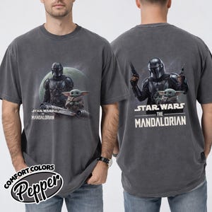 May include: Dark gray t-shirt featuring a graphic of The Mandalorian and Grogu from Star Wars. The front shows the characters with a spaceship and a planet in the background. The back of the shirt shows the Mandalorian holding blasters with Grogu.