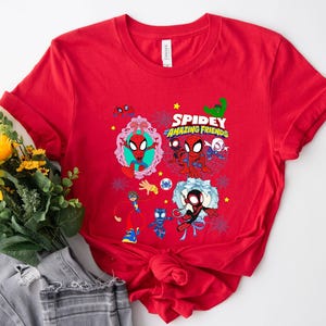 May include: Red t-shirt featuring a colorful graphic of Spidey and his Amazing Friends. The design includes various characters and the text "Spidey and Amazing Friends". The shirt is made of a soft material.