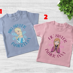 May include: Two children's t-shirts. One is light blue with the text "Big Sister I'm Her Elsa" and a graphic of Elsa. The other is pink with "Lil' Sister I'm Her Anna" and a graphic of Anna.