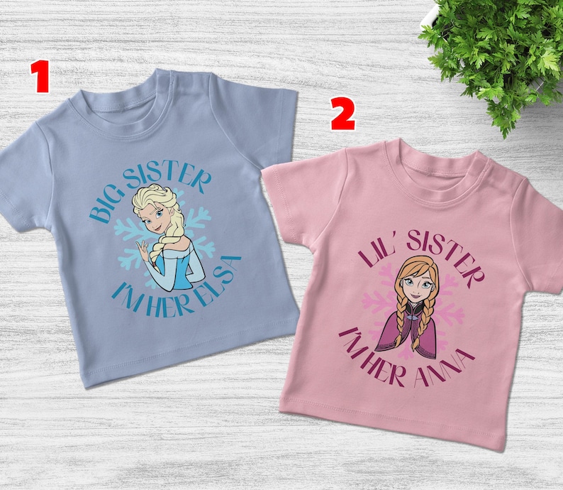 May include: Two children's t-shirts. One is light blue with the text "Big Sister I'm Her Elsa" and a graphic of Elsa. The other is pink with "Lil' Sister I'm Her Anna" and a graphic of Anna.