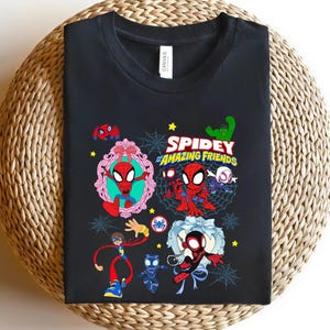 May include: Black t-shirt featuring a colorful graphic of Spider-Man and his friends. The design includes the text "Spidey and Amazing Friends" with various cartoon characters and web designs. The shirt is displayed on a woven surface.
