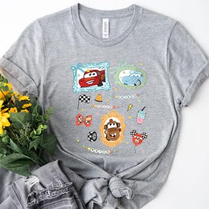 Puede incluir: Una camiseta gris con un dise&ntilde;o que presenta im&aacute;genes de coches de dibujos animados enmarcadas y otros gr&aacute;ficos. El dise&ntilde;o incluye un coche rojo, un coche azul, una bandera a cuadros y un batido. La palabra "Mater" tambi&eacute;n es visible.
