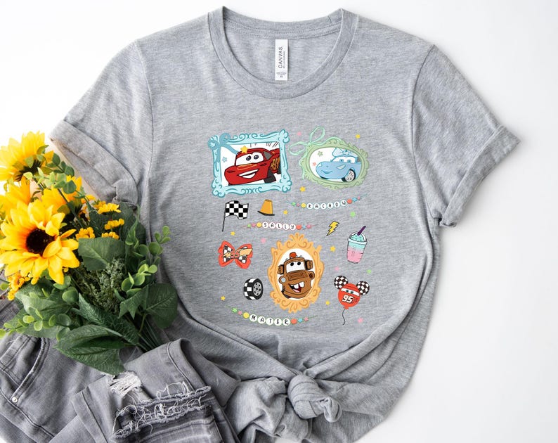 Puede incluir: Una camiseta gris con un dise&ntilde;o que presenta im&aacute;genes de coches de dibujos animados enmarcadas y otros gr&aacute;ficos. El dise&ntilde;o incluye un coche rojo, un coche azul, una bandera a cuadros y un batido. La palabra "Mater" tambi&eacute;n es visible.