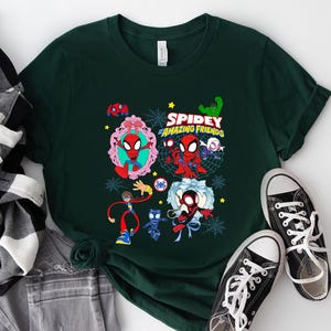 May include: Dark green t-shirt featuring a colorful graphic of Spider-Man and friends with the text "Spidey and His Amazing Friends". The design includes various characters and spiderwebs. Black canvas sneakers with white laces are next to the shirt.
