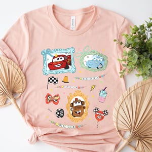 Puede incluir: Una camiseta rosa claro con im&aacute;genes de dibujos animados de personajes de la pel&iacute;cula Cars. El dise&ntilde;o incluye im&aacute;genes enmarcadas de Rayo McQueen, Sally y Mate, junto con otros gr&aacute;ficos tem&aacute;ticos y los nombres "Sally" y "Mater".