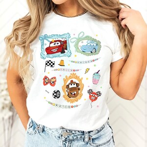 Puede incluir: Camiseta blanca con im&aacute;genes de dibujos animados de personajes de Cars, incluyendo a Rayo McQueen y Mate, en marcos decorativos. Otros gr&aacute;ficos incluyen una bandera a cuadros, un cono de tr&aacute;fico y un globo.