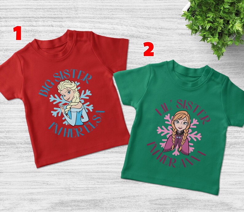 May include: Two short-sleeved t-shirts, one red and one green. The red shirt features a graphic of Elsa with the text "Big Sister" and "Father Elsa". The green shirt has a graphic of Anna with the text "Lil' Sister" and "Father Anna".