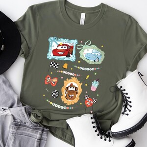Puede incluir: Camiseta verde oliva con personajes de coches de dibujos animados enmarcados. El dise&ntilde;o incluye un coche rojo, un coche azul y un coche marr&oacute;n, junto con elementos decorativos como banderas a cuadros, estrellas y un batido. La camiseta se combina con botas blancas y vaqueros.