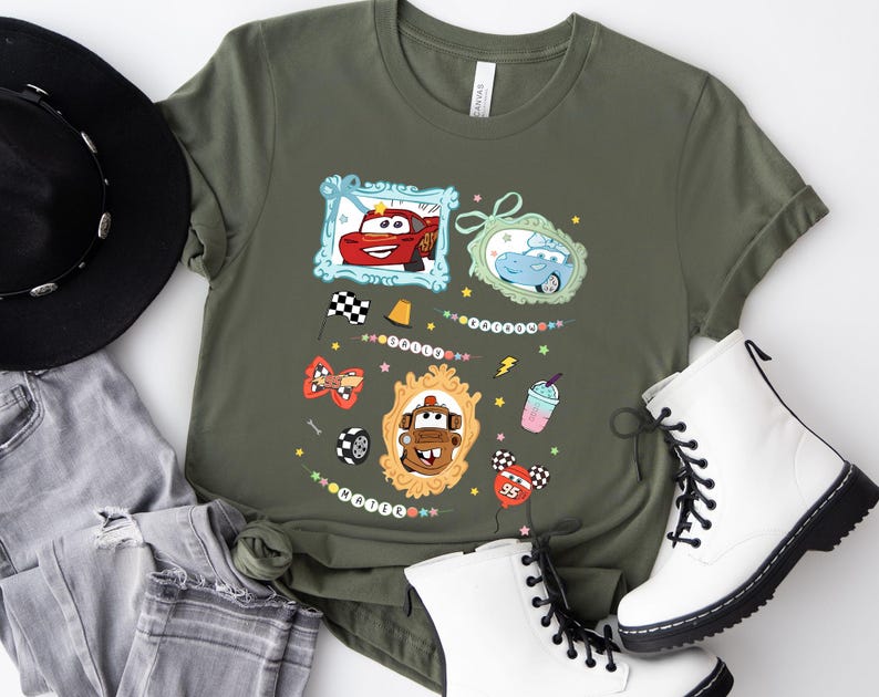 Puede incluir: Camiseta verde oliva con personajes de coches de dibujos animados enmarcados. El dise&ntilde;o incluye un coche rojo, un coche azul y un coche marr&oacute;n, junto con elementos decorativos como banderas a cuadros, estrellas y un batido. La camiseta se combina con botas blancas y vaqueros.