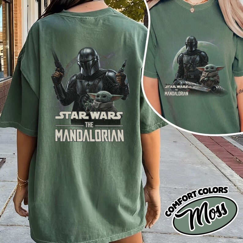 May include: Green t-shirt featuring a graphic of The Mandalorian and Grogu from Star Wars. The design includes the text "STAR WARS THE MANDALORIAN". The shirt is a comfort color in the color "Moss".