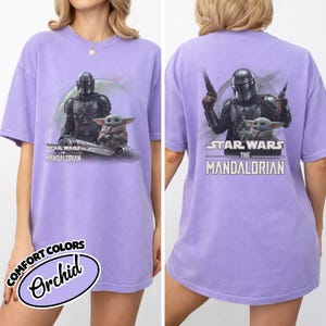 May include: Orchid-colored t-shirt featuring a graphic of The Mandalorian and Grogu from Star Wars. The front shows the characters with the text "STAR WARS THE MANDALORIAN". The back of the shirt also displays the characters and text. The shirt is a Comfort Colors brand.