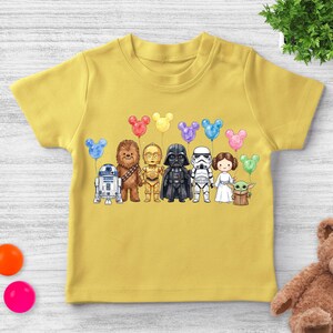 Op de afbeelding: Mosterdgeel T-shirt met korte mouwen met een cartoonillustratie van Star Wars-personages die Mickey Mouse-vormige ballonnen vasthouden. De personages zijn R2-D2, Chewbacca, C-3PO, Darth Vader, een Stormtrooper, Princess Leia en Grogu.
