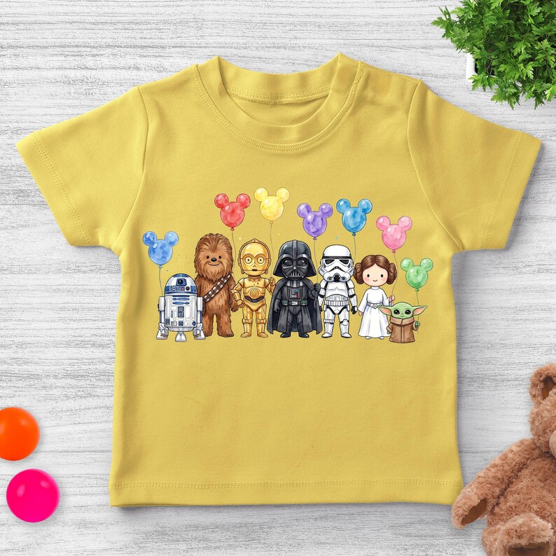 Op de afbeelding: Mosterdgeel T-shirt met korte mouwen met een cartoonillustratie van Star Wars-personages die Mickey Mouse-vormige ballonnen vasthouden. De personages zijn R2-D2, Chewbacca, C-3PO, Darth Vader, een Stormtrooper, Princess Leia en Grogu.