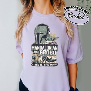 Puede incluir: Una camiseta de color lila claro con un gr&aacute;fico de The Mandalorian y Grogu de Star Wars. El dise&ntilde;o incluye el casco del Mandaloriano, Grogu y el texto "THE MANDALORIAN AND GROGU" y "THIS IS THE WAY". La camiseta tambi&eacute;n tiene el texto "COMFORT COLORS" y "Orchid".