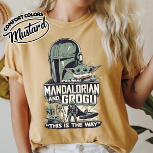 Puede incluir: Camiseta de color mostaza con un gr&aacute;fico de Star Wars de The Mandalorian y Grogu. El dise&ntilde;o incluye el casco del Mandaloriano, Grogu y la frase "THIS IS THE WAY". La camiseta tambi&eacute;n tiene las palabras "COMFORT COLORS" y "Mustard".