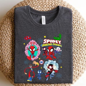 May include: Dark gray t-shirt featuring a colorful graphic of Spider-Man and friends. The design includes the text "Spidey and Amazing Friends" with various cartoon illustrations of the characters. The shirt is folded on a woven surface.