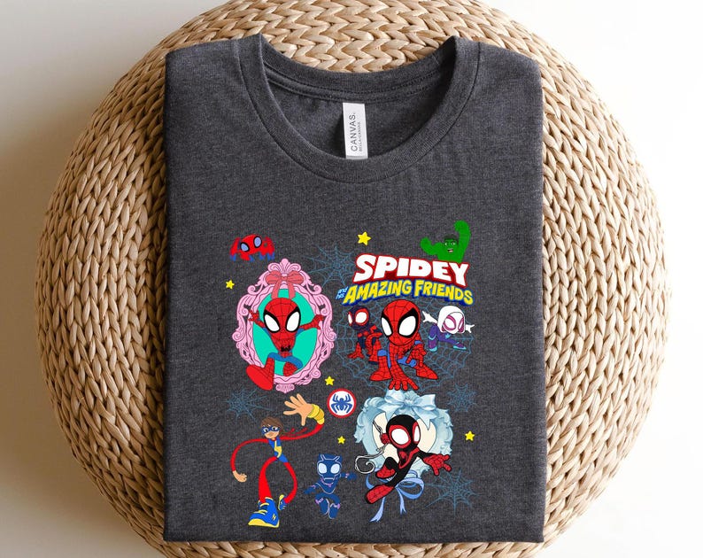 May include: Dark gray t-shirt featuring a colorful graphic of Spider-Man and friends. The design includes the text "Spidey and Amazing Friends" with various cartoon illustrations of the characters. The shirt is folded on a woven surface.