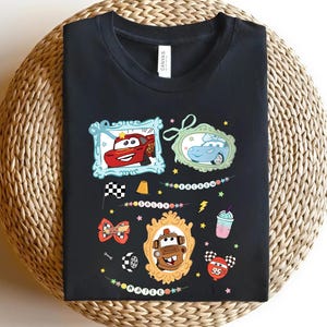 Puede incluir: Camiseta negra con gr&aacute;ficos de coches de dibujos animados coloridos. El dise&ntilde;o incluye im&aacute;genes enmarcadas de un coche de carreras rojo, un coche azul y un cami&oacute;n marr&oacute;n. Elementos adicionales: bandera a cuadros, rayo y batido.