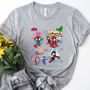 May include: A gray t-shirt featuring the text "Spidey and His Amazing Friends" with cartoon illustrations of Spider-Man and other characters. The design includes various colorful elements and is printed on a soft, casual tee.