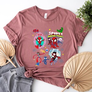May include: A dusty rose t-shirt featuring a colorful graphic of Spider-Man and his friends. The design includes the text "Spidey and His Amazing Friends" with various cartoon characters. The shirt is displayed with folded sleeves and a casual style.