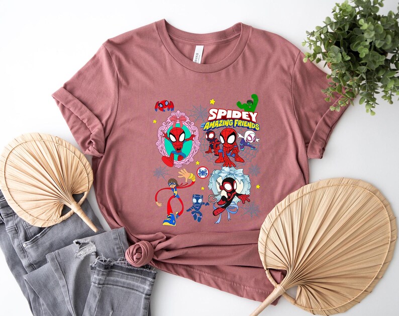 May include: A dusty rose t-shirt featuring a colorful graphic of Spider-Man and his friends. The design includes the text "Spidey and His Amazing Friends" with various cartoon characters. The shirt is displayed with folded sleeves and a casual style.