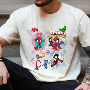 May include: Cream-colored t-shirt featuring a colorful graphic of Spider-Man and friends. The design includes the text "Spidey and His Amazing Friends" and various cartoon illustrations of the characters. The shirt is short-sleeved.