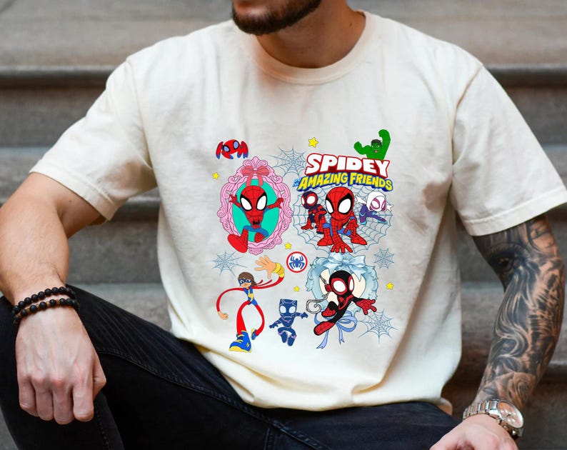 May include: Cream-colored t-shirt featuring a colorful graphic of Spider-Man and friends. The design includes the text "Spidey and His Amazing Friends" and various cartoon illustrations of the characters. The shirt is short-sleeved.