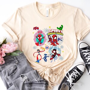 May include: A beige t-shirt featuring a colorful graphic of Spider-Man and friends. The design includes the text "Spidey Amazing Friends" and various cartoon characters. The shirt is paired with gray jeans and black sneakers.