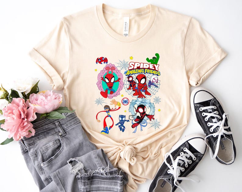 May include: A beige t-shirt featuring a colorful graphic of Spider-Man and friends. The design includes the text "Spidey Amazing Friends" and various cartoon characters. The shirt is paired with gray jeans and black sneakers.
