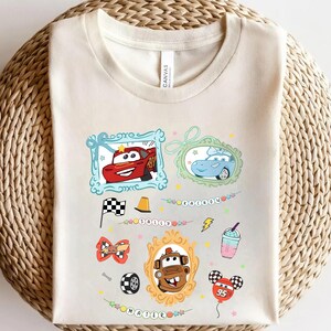 Puede incluir: Camiseta color crema con im&aacute;genes de dibujos animados de coches en marcos, junto con otros gr&aacute;ficos como una bandera a cuadros, un cono de tr&aacute;fico y un globo. La camiseta tiene la palabra "CARS" y el n&uacute;mero "95".