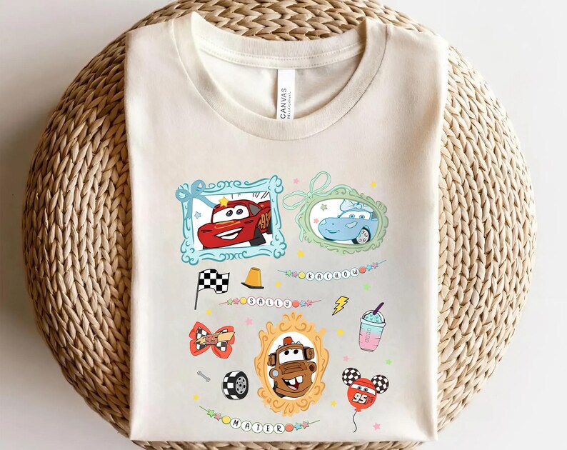 Puede incluir: Camiseta color crema con im&aacute;genes de dibujos animados de coches en marcos, junto con otros gr&aacute;ficos como una bandera a cuadros, un cono de tr&aacute;fico y un globo. La camiseta tiene la palabra "CARS" y el n&uacute;mero "95".