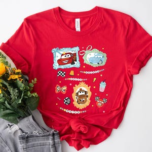 Puede incluir: Camiseta roja con personajes de dibujos animados de coches en marcos ornamentados, banderas de carreras, un rayo y una bebida. El dise&ntilde;o incluye el texto "Cars".