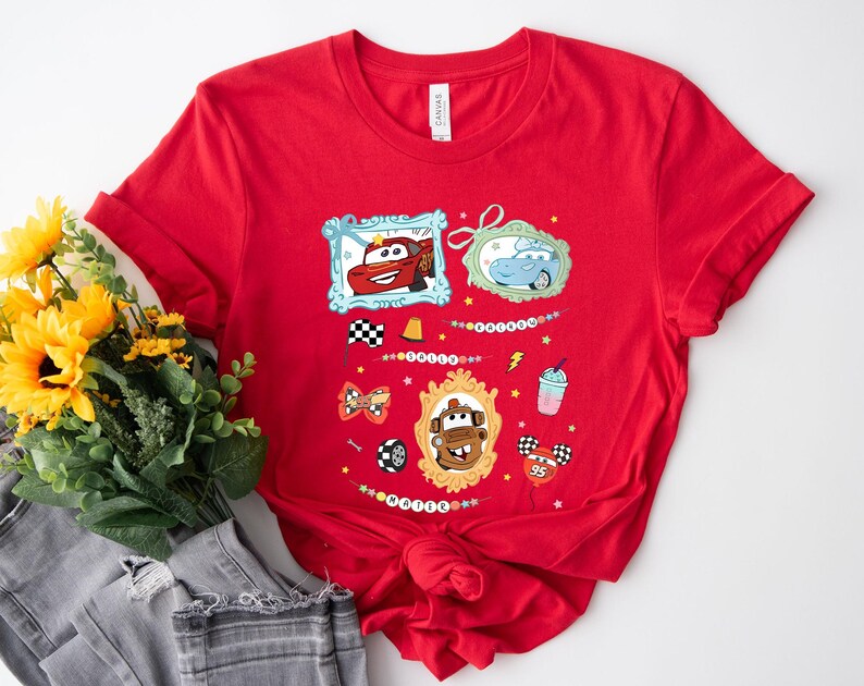 Puede incluir: Camiseta roja con personajes de dibujos animados de coches en marcos ornamentados, banderas de carreras, un rayo y una bebida. El dise&ntilde;o incluye el texto "Cars".