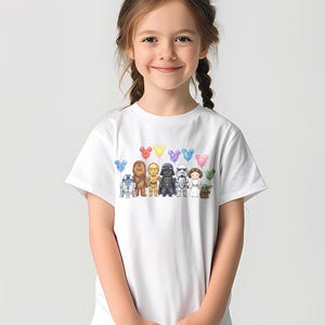 Op de afbeelding: Wit T-shirt met een kleurrijke illustratie van Star Wars-personages en Mickey Mouse-vormige ballonnen. Het ontwerp bevat R2-D2, Chewbacca, C-3PO, Darth Vader, een Stormtrooper, Princess Leia en Grogu.