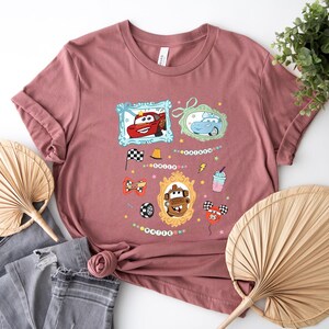Puede incluir: Camiseta rosa polvoriento con personajes de dibujos animados de coches en marcos ornamentados. Gr&aacute;ficos adicionales incluyen una bandera a cuadros, un neum&aacute;tico de carreras y un batido. La camiseta tiene cuello redondo y mangas cortas.
