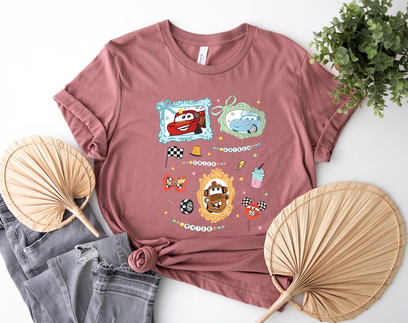Puede incluir: Camiseta rosa polvoriento con personajes de dibujos animados de coches en marcos ornamentados. Gr&aacute;ficos adicionales incluyen una bandera a cuadros, un neum&aacute;tico de carreras y un batido. La camiseta tiene cuello redondo y mangas cortas.