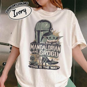 Puede incluir: Camiseta de color marfil con un gr&aacute;fico de The Mandalorian y Grogu de Star Wars. El dise&ntilde;o incluye el casco del Mandaloriano, Grogu y el texto "THIS IS THE WAY". La camiseta tambi&eacute;n tiene el texto "COMFORT COLORS" e "Ivory".