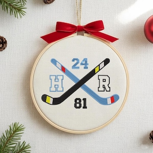 May include: A hockey-themed embroidery hoop ornament. The design features crossed hockey sticks, the numbers 24 and 81, and the letters H and R. A red bow and gold string are at the top for hanging.