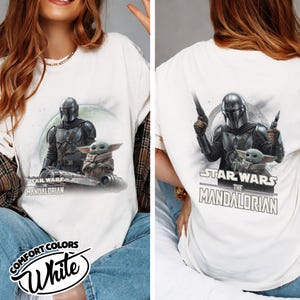 May include: White t-shirt featuring a graphic of The Mandalorian and Grogu from Star Wars. The front shows the characters on a vehicle, while the back displays them with blasters. The shirt includes the text "STAR WARS THE MANDALORIAN".