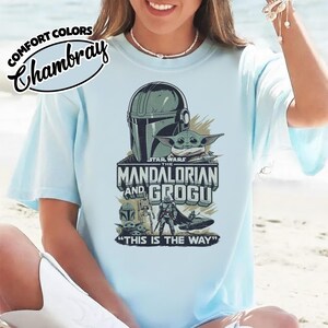 Puede incluir: Camiseta azul claro con un dise&ntilde;o de Star Wars de The Mandalorian y Grogu. El dise&ntilde;o incluye el casco del Mandaloriano, Grogu y el texto "THE MANDALORIAN AND GROGU" y "THIS IS THE WAY". La camiseta tiene la inscripci&oacute;n "COMFORT COLORS Chambray".
