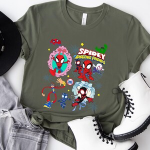 May include: Olive green t-shirt featuring a colorful graphic of Spidey and his Amazing Friends. The design includes various characters and the show's logo. The shirt is paired with white boots, a black hat, and gray jeans.