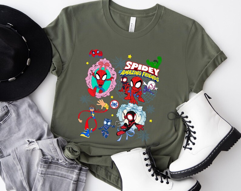 May include: Olive green t-shirt featuring a colorful graphic of Spidey and his Amazing Friends. The design includes various characters and the show's logo. The shirt is paired with white boots, a black hat, and gray jeans.