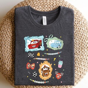 Puede incluir: Camiseta gris oscuro con personajes de coches de dibujos animados en marcos decorativos. El dise&ntilde;o incluye un coche rojo, un coche azul y un coche marr&oacute;n, adem&aacute;s de una bandera a cuadros y un batido. La camiseta es de material suave.