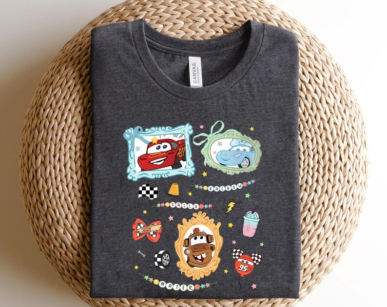 Puede incluir: Camiseta gris oscuro con personajes de coches de dibujos animados en marcos decorativos. El dise&ntilde;o incluye un coche rojo, un coche azul y un coche marr&oacute;n, adem&aacute;s de una bandera a cuadros y un batido. La camiseta es de material suave.