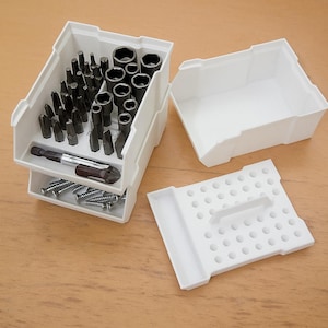 Può includere: Un organizer per utensili in plastica bianca con più scomparti. L'organizer contiene varie punte per cacciavite nere, prese e viti. Sono presenti anche un coperchio e un contenitore vuoto, che suggeriscono un set per organizzare utensili e hardware.